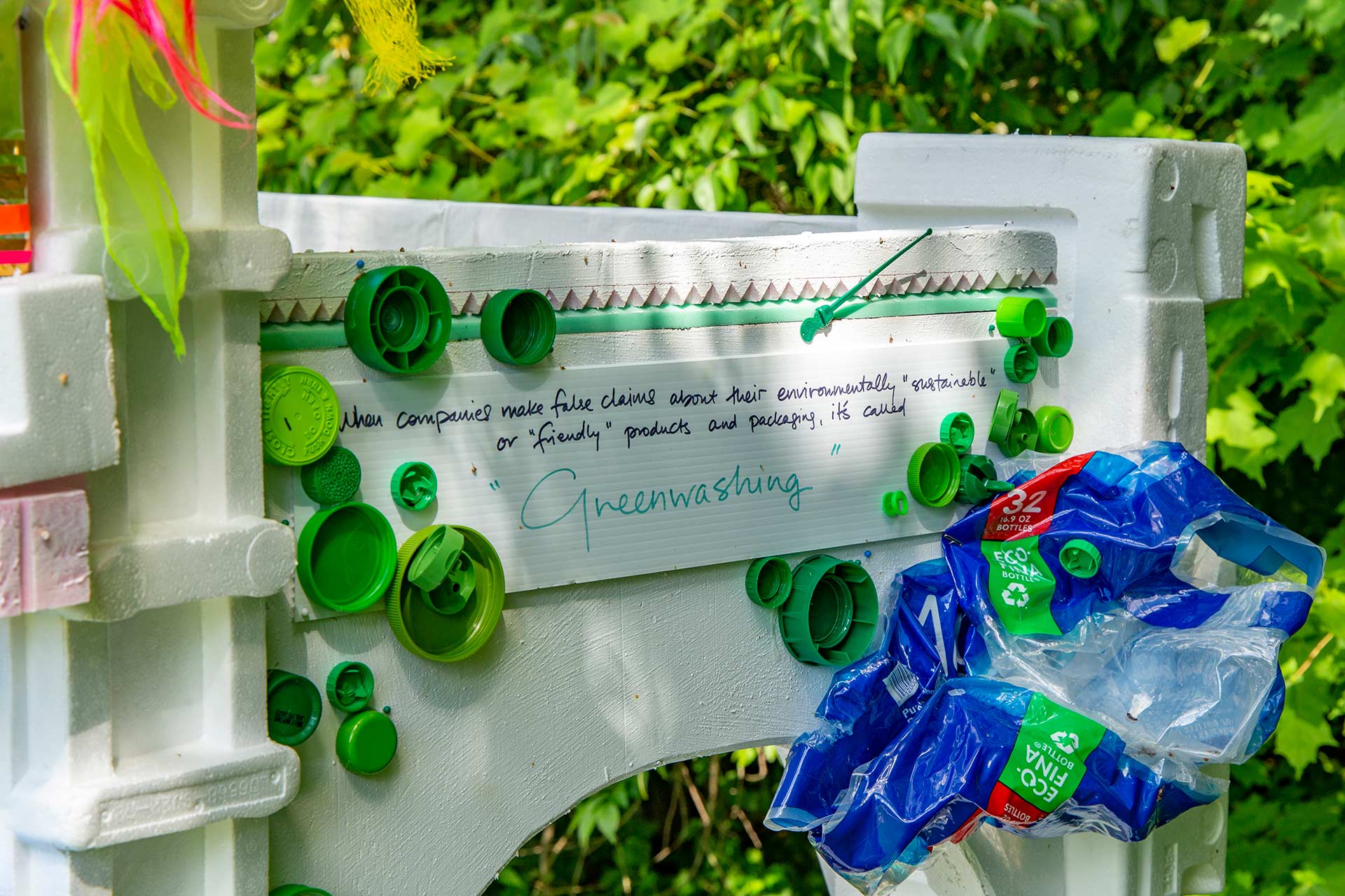 Many green bottle caps and a plastic cover for bottled waters glued to a white arch doorway that has a sentence about 'Greenwashing'