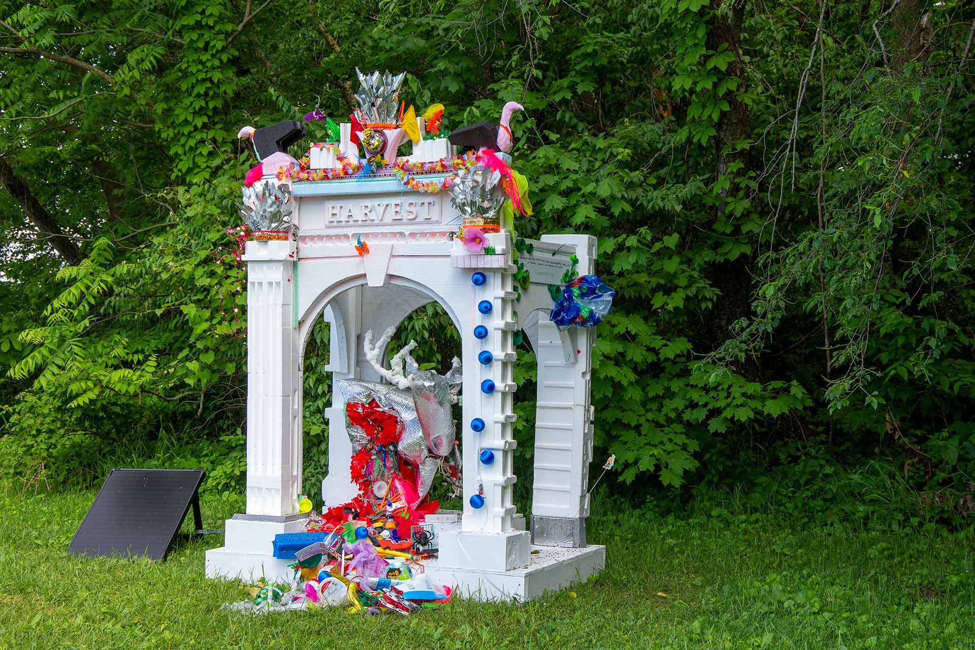 A white mini pavilion with arch doorways on the side that has colorful decoration and the word 'harvest' on it