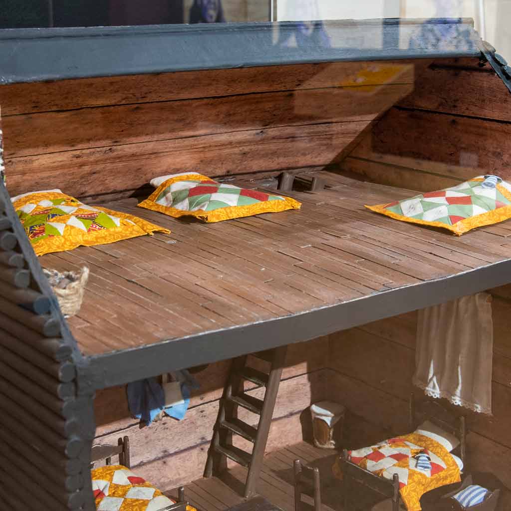 A wooden dollhouse with two floors showing multiple beds with yellow, white, green, and red quilts