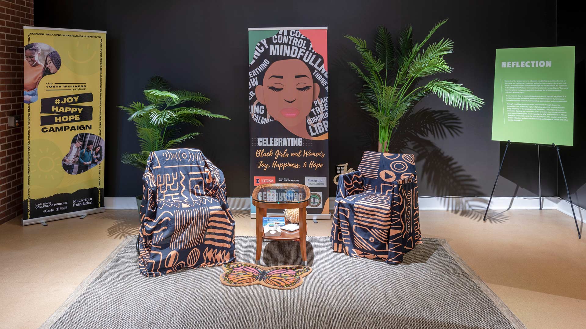 An overview of the reflection section of the exhibit with two chairs, a table, and a butterfly rug on top of a large gray rug