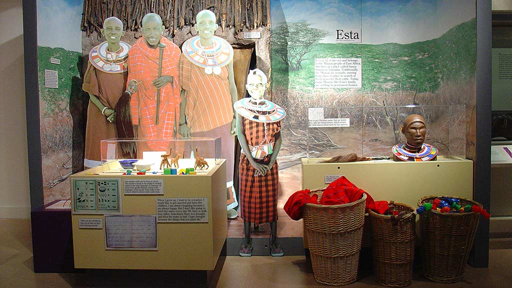 children in Esta section, girl wearing a long dress with various figures in a display case to her right and other objects in baskets to her left