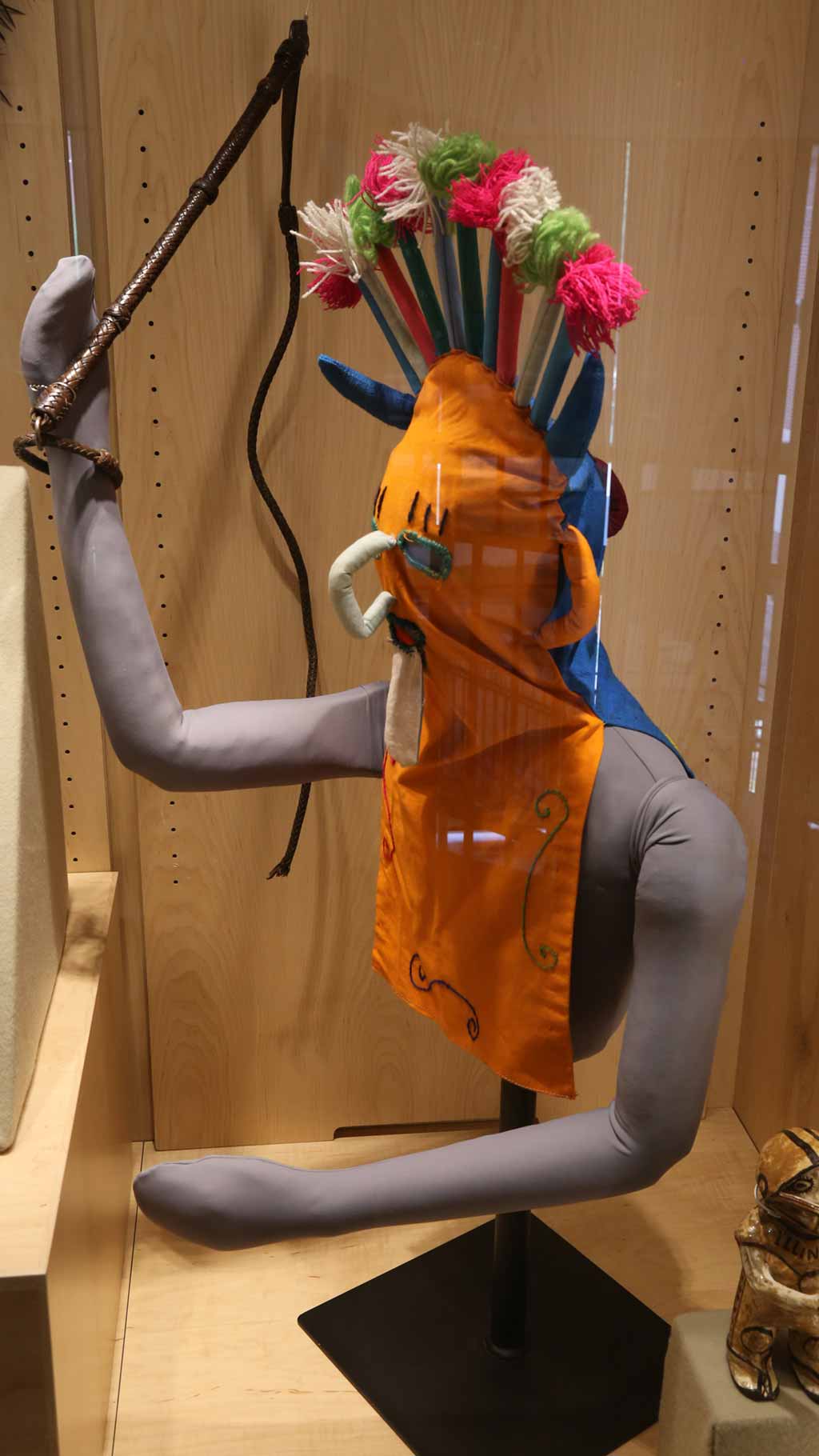 mannequin holding whip and wearing an orange face covering that has some designs on top