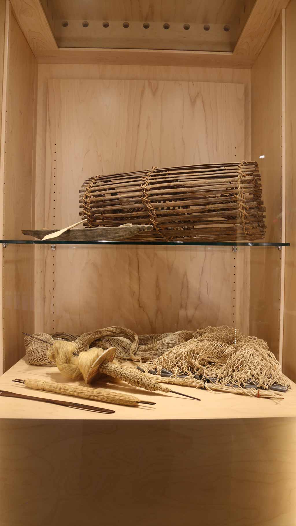 vertical shot of the exhibit case with the fish net, shuttles, fish trap and canoe